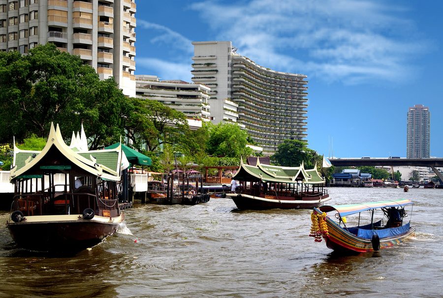 A passenger boat on the Chao Phraya River with Bangkok buildings along the shore.